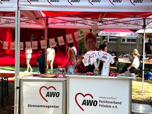 AWO Info-Stand. Rot-Weißer Pavillon. Eine Frau steht darin. Info-Material, Fähnchen, Schlüsselbänder gibt es dort.