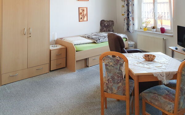 Bewohnerzimmer- Links an der Wand ein Kleiderschrank, Nachttisch und Bett. Rechts: ein Fenster, ein Fernsehtischen mit TV und im Raum noch ein kleiner Tisch mit zwei Stühlen.