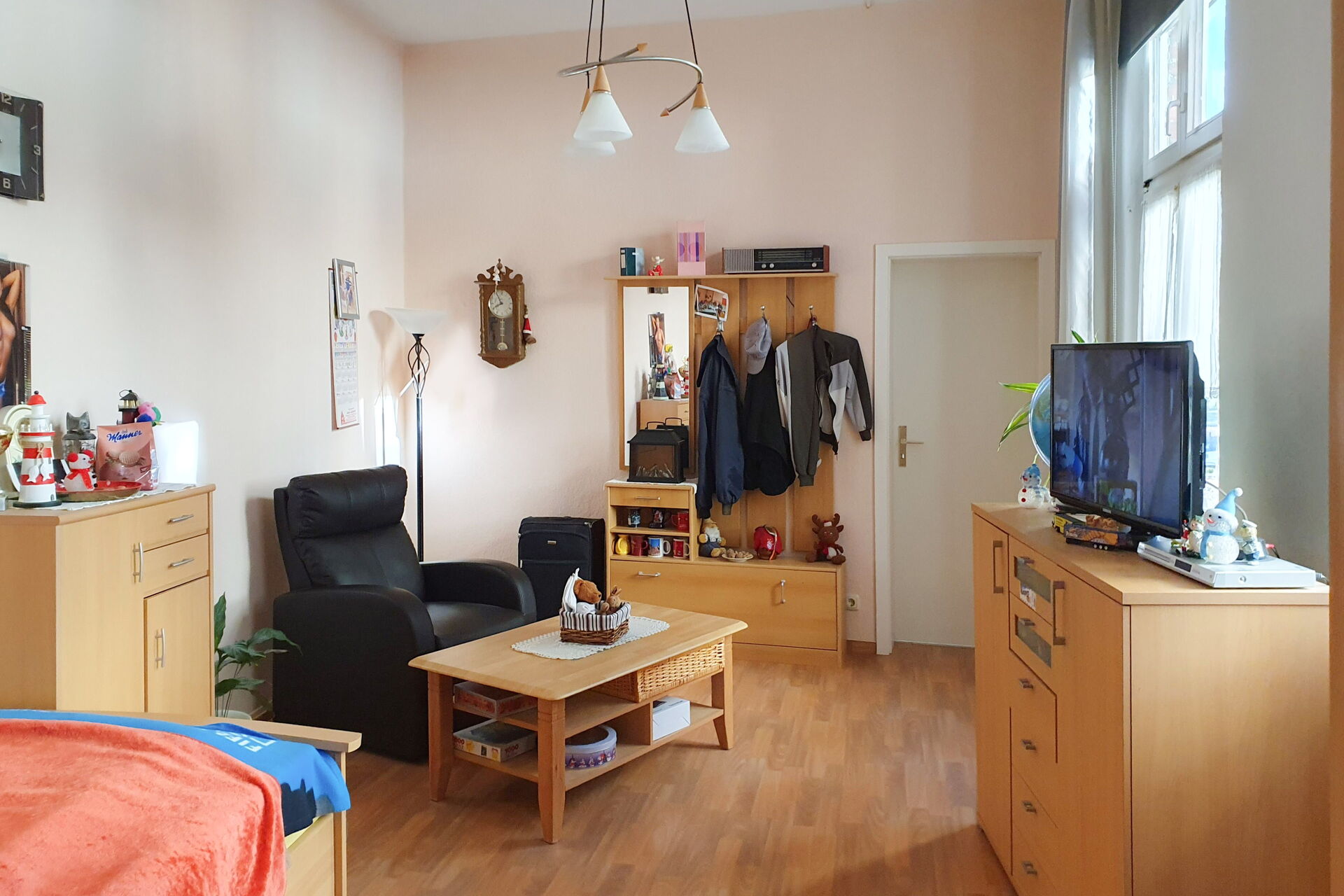 Bewohnerzimmer. Bick durch dem Raum zur Tür. Neben der Tür Garderobe, ein schwarzer Fernsehsessel, davor ein kleiner Couchtisch. Bewohntes Zimmer mit Kleidung und Deko.
