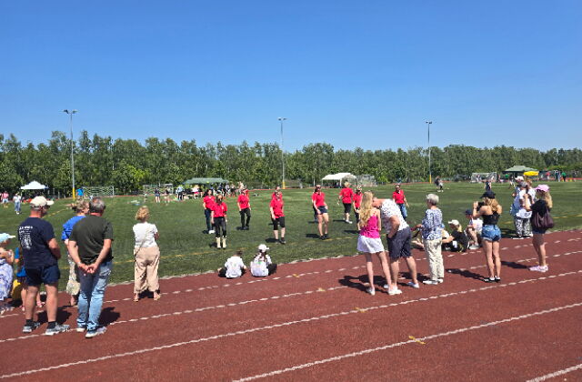 Ein Sportplatz mit Laufstrecke und Grün. Jugendliche in roten Sporttrikots auf der Grünfläche.