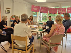 Raum, Tisch. 8 Personen sitzen um den Tisch, Hinten im Raum hängen rosa Schilder mit Forderungen Text: Mehr Qualität in katas und Schulen.