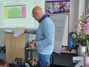 Mann erklärt Präsentation die an der Wand läuft. Auf dem Fensterbrett rechts stehen Orchideen.