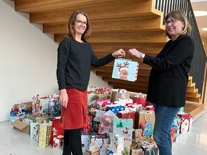 Unter einen freien Treppe stehen sehr viele bbunte Geschenktüten. Zwei Frauen stehen davor lächeln und halten gemeinsam eine kleine Geschenktüte mit Rudolf als Bild darauf.
