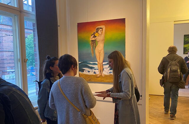 Ausstellungsraum. Drei Frauen stehen vor einem Bild an der Wand. Das Bild zeigt eine nackte Frau mit langen Haaren am Strand stehend. Die Hintergrund ist Regenbogen-farbig.