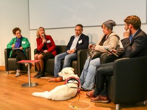 5 Personen und ein Hund. 3 Frauen und zwei Männer sitzen in einer Gesprächsrunde. Frau im roten Blazer hat ein Mikro in der Hand. Vor einer Frau mit Mütze liegt ein weißer Blindenhund.