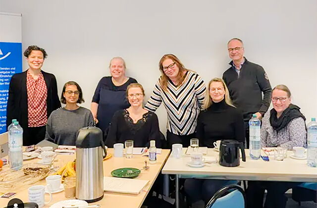In einem Raum. Tisch gedeckt mit Kaffee, Wasser. 4 Personen sitzen am Tisch, 4 Personen stehen versetzt dahinter. 7 Frauen, ein Mann, alle lächeln in die Kamera.