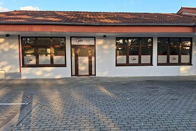 gepflasterte Parkfläche mit grauen Steinen, im Hintergrund ein Gebäude mit einer weißen Fassade, einem roten Dach, mit einer Glastür mittig, links von der Glastür eine Fensterglasfront, rechts der Glastür zwei Fensterglasfronten
