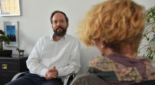 Ein Büroraum. Mann sitzt einer Frau gegenüber. Die Kamera ist auf den Mann gerichtet, die Frau nur von hinten im Bild. Mann trägt weißes langärmliches Hemd, hat einen Vollbart und dunkle Haare.