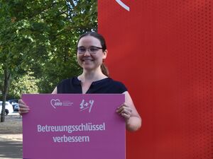 Portrait: Junge Frau mit braunen langen Haaren, Mittelscheitel und Zopf, sie trägt ein schwarzes T-Shirt und lächelt. Sie hält ein pinkes Schild vor sich mit Text: Betreuungsschlüssel verbessern.