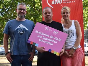 Portrait: Großer Mann, graue Haare, Vollbart, Brille, T-Shirt, kurze blaue Jeans, Mann in schwarzer, kurzer Kleidung, blond und eine Frau mit langen blonden Haaren, weißem Träger-Shirt und lachsfarbener leichten Stoffhose stehen draußen.