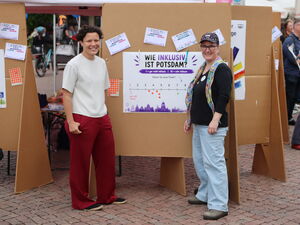 2 Frauen vor einer Stellwand mit Plakat. Die Frage: Wie inklusiv ist Potsdam?   Darunter ein Strahl von 1 bis 10.