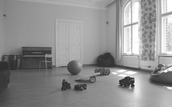 Großer Raum mit großen Fenstern. In dem Raum, ein Sitzsack in der Ecke, 2 Paar Rollschuhe, ein Sitzball, ein Klavier mit Stuhl davor.