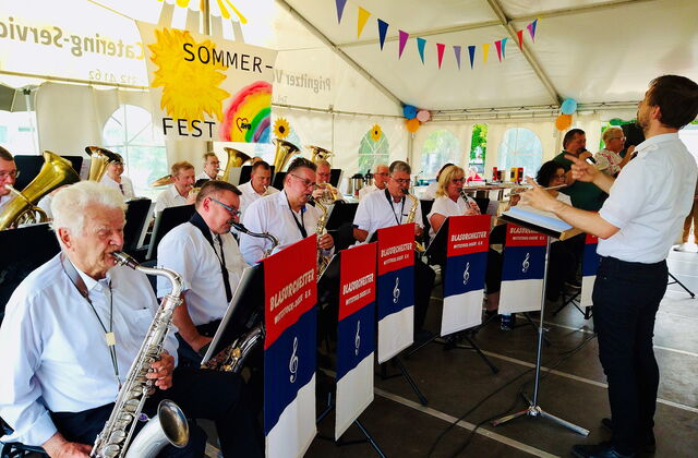 Musiker mit Blasinstrumenten und rechts der Dirigent, alle tragen weiße Hemden uns schwarze Hosen.