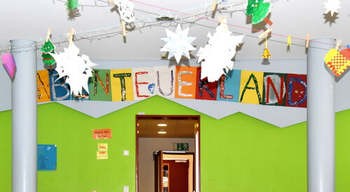 Ein Raum mit offener Tür. Die Tür mittig. Zwei graue Säulen im Raum. Die Wand ist grün. Über der Tür farbige A4 Seiten mit bunten Groß-Buchstaben Wort: ABENTEUERLAND