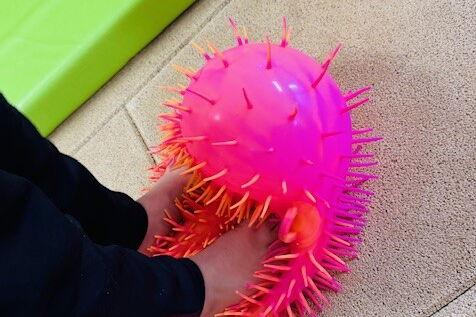 Ein Kind steht mit nackten Füßen auf einem pinken, weichen "Igelball". Am Bildrand ist eine grüne Matte zu sehen.