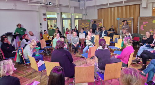 Großer Raum, Gesprächsrunde Menschen sitzen in Stuhlkreisen. Hinten im Raum sind 4 Flipcharts an denen farbige Moderationskarten angepinnt sind.