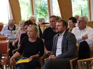 Mehrere Menschen sitzen in einem großen Raum. In der ersten Reihe, eine blonde Frau mit Brille auf den Kopf geschoben, die hat einen gelben Hefter auf dem Schoß. Daneben ein Mann mit weißen Hemd und grauen Anzug.