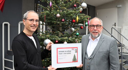 Zwei Männer um die 50 mit einer eingerahmten Spende. Text: Spende in Höhe von 1.100 Euro. Die Männer stehen draußen vor einen bunt geschmückten Weihnachtsbaum. Beide graue Haare, Brille, sind feierlich gekleidet grauer Anzug / weißes Hemd.