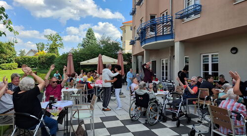 Eine große Terrasse, Stühle, Tische, Menschen in Rollstühlen und mit Rollatoren. Es herrscht ausgelasssene Stimmung, einige schwenken die Arme in der Luft und in der Mitte wird getanzt.