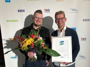 Zwei Männer vor einer Werbewand. Der jüngere Mann hat einem großen Blumenstrauß in der rechten Hand. Der Mann daneben hält eine gerahmte Urkunde zum 3. Platz vor sich.