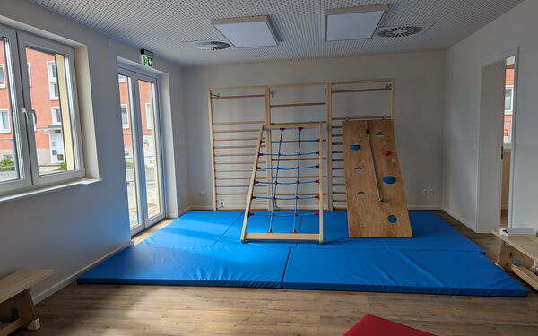 Sportraum. Große blaue Matten liegen vor eine Sprossenwand. An der Sprossenwand lehnen schräg zum rauf klettern zwei Kletterbretter.
