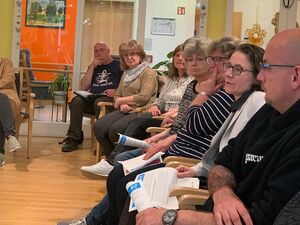 Großer Raum. 8 Menschen sitzen in einer Reihe. Stuhlreihe von der Seite fotografiert.