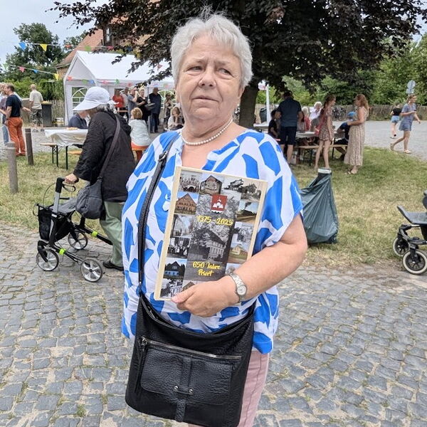 Eine Frau im freien, hinter ihr kleines Fest. Sie hält eine Art Proschüre in die Kamera mit historischen Fotos und dem Text: 1375 - 2025   650 Jahre Priort