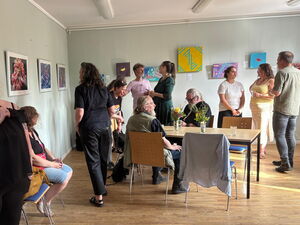 Ausstellung. Bilder hängen an weißen Wänden. Menschen sehen sich die Bilder an.
