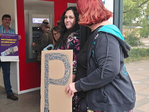 Menschen-Schlange vor Geldautomat. Einige Frauen halten ein großes P In die Kamera. Ein Mann steht links außen und hält ein lila Plakat "Wie komme ich an mein Geld?"