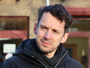Ein Mann mit dunklen Haaren, etwas Bart und dunkler Kleidung steht vor einem Gebäude.