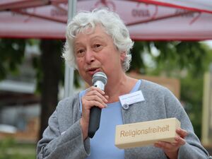 Ältere Frau mit Mikro in der rechten Hand und einem Holzklotz mit dem Wort: Religionsfreiheit. Frau hat weiße gelockte Haare, eine hellgraue Strickjacke über einem hellblauen Oberteil. Sie spricht in das Mikrofon.