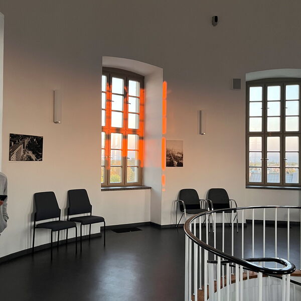 Ein leerer Flur mit Stühlen an der Wand, orangenes Sonnenlicht (Abend) fällt durch ein großes Fenster.