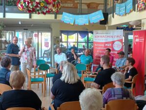 Großer heller Raum, viele Stühle in Kreisen angeordnet. Menschen sitzen auf den Stühlen. In der Mitte stehen drei Menschen, zwei Frauen ein Mann. Eine Frau hält ein Mikrofon in der Hand.