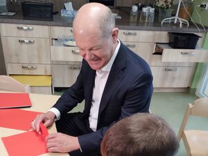 Ein kleiner Junge sitzt an einem kleinen Tisch und schließt einen roten Umschlag. Ein Mann im dunklen Anzug und weißem Hemd sitzt neben ihm und tut es ihm nach. Der Mann lächelt.