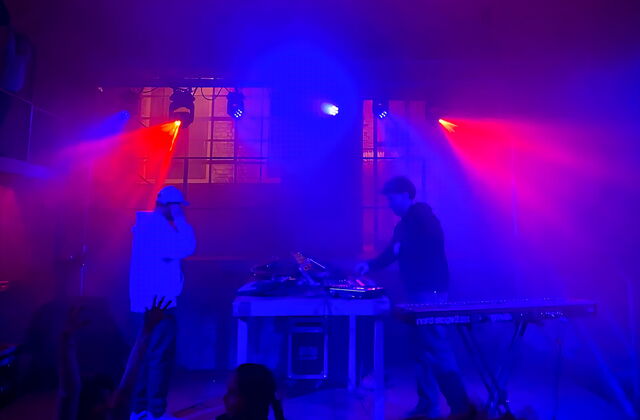 DJs am Pult, es ist dunkel, rot blaues Scheinwerferlicht sorgt für Party-Feeling. Tanzende Menschen im Vordergrund.
