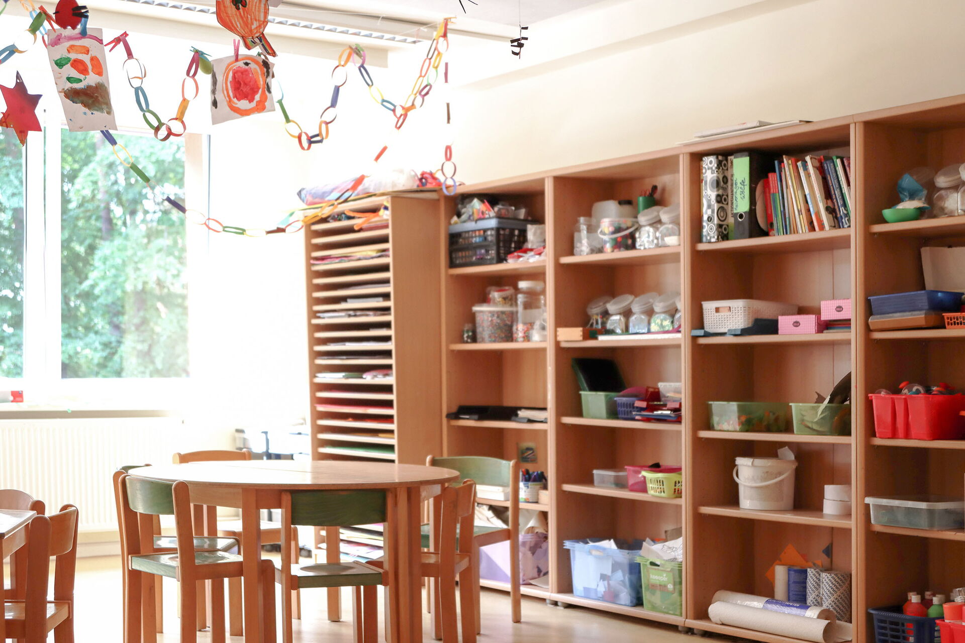Großes Wandregal mit Bastelmaterialien. Ein Runder Tisch mit Stühlen. Von der Decke hängen gebastelte Strene und Girlanden und Bilder von Kindern.