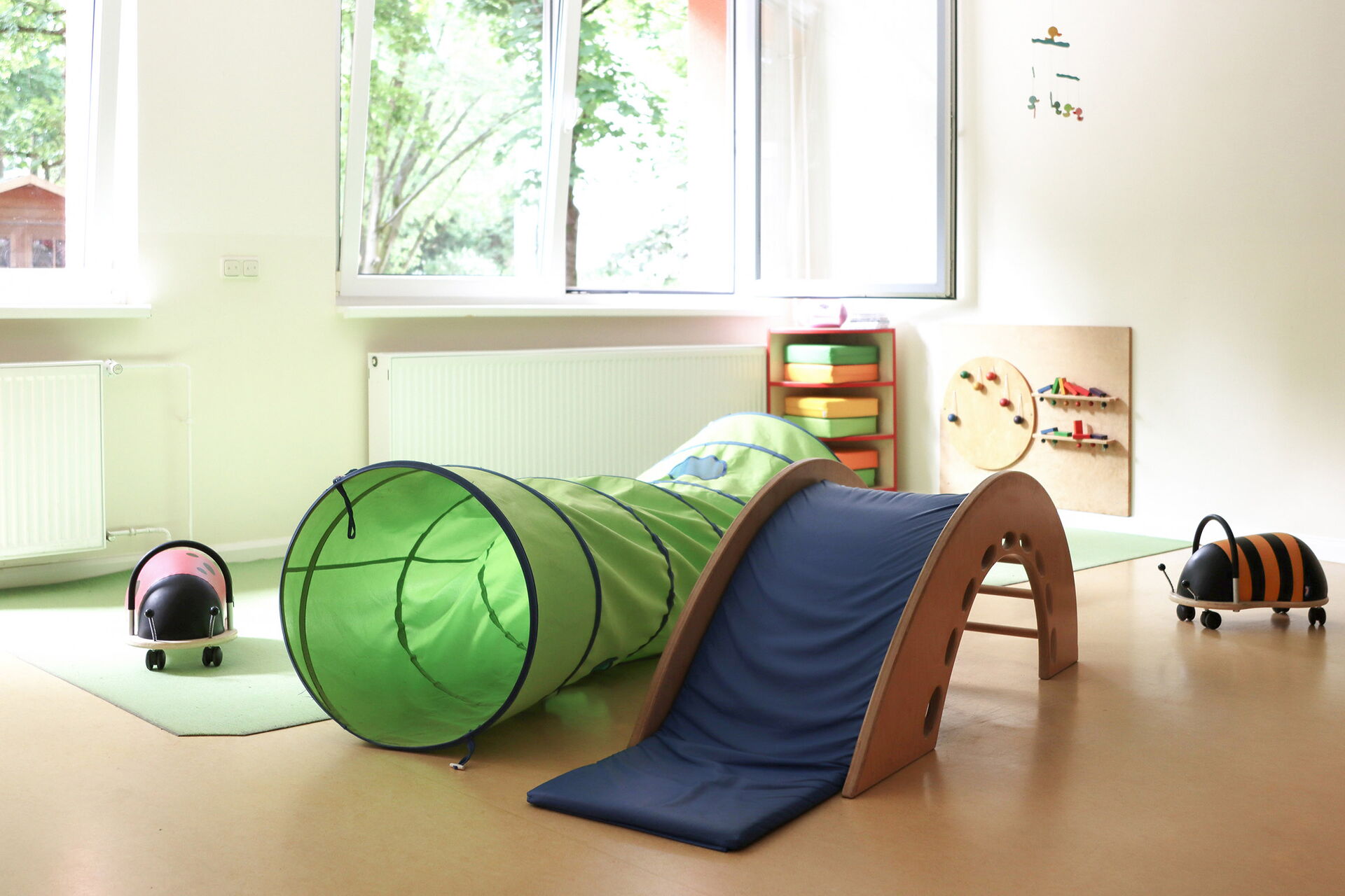 Helles Spielzimmer. Im Vordergrund befinden sich ein grüner Kriechtunnel, eine kleine blaue Rutsche aus Holz und zwei Spielzeuge, die wie Käfer aussehen.