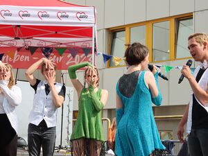 Tanzgruppe aus jugendlichen Mädchen und Jungs. Mädchen im blauen Kleid und Junge im schwarzen T-Shirt und weißer Weste stehen sich gegenüber und singen. 3 weitere Tänzer performen im Hintergrund.