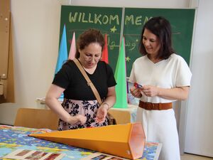 Zwei Frauen in einem Klassenraum, die eine gelbe Zuckertüte bunt bekleben. An der Schultafel steht WILLKOMMEN und hinter den Frauen stehen weitere farbige Zuckertüten.