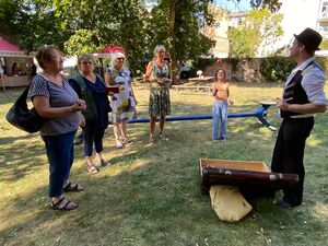 Mann mit Hut, weißem Hemd und schwarzer Weste und Hose steht vor einem geöffneten Koffee. Drumherum stehen vier Frauen und schauen erwartungsvoll.. Ein Mädchen wirf einen roten Ball hoch.