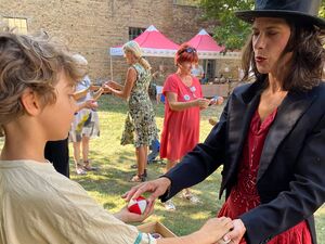 Frau mit Zylinder, roten Kleid darüber ein scharzer Bolero, Ihr gegenüber steht ein Junge, Sie hat zwei Bälle, einen rot-weißen Ball hat der Junge in der linken Hand, den anderen hält die Frau und der Junge hat die Hand darüber gelegt.