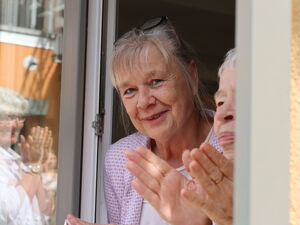 Frau blickt aus einem Fenster in die Kamera und lächelt. Am Fenter neben ihr eine Person die Klatscht.