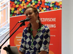 Frau auf einem Hocker, Mikrofonständer vor ihr. Hinter ihr rote Roll-Ups der Frühförderstelle.