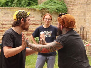 Draußen auf Wiese: Workshop: 2 Männer in der Mitte stehen sich Handfläche an Handfäche und drücken sich weg. Im Kreis stehen Frauen ringsum.
