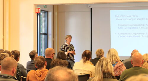 In einem Raum sitzen Menschen. Von hinten fotografiert. Vorn an der Leinwand eine Präsentation über Förderrichtlinie Klimaanpassung in sozialen Einrichtungen.