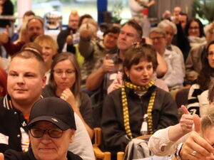 Voller Saal mit Teilnehmenden. Ein Mittelgang ist frei geräumt. Einige der Teilnehmenden lächeln und strecken den Daumen-Hoch.