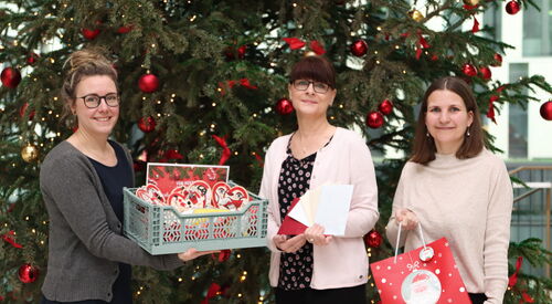 Drei Frauen vor einem großem geschmücktem Weihnachtsbaum mit roten Kugeln und Schleifen. Zwei Frauen tagen eine Faltbox mit Herzen drin. Die dritte hat eine rote Weihnachtstüte mit Aufschrift: Best Wishest