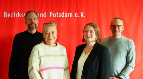 Gruppenfoto vor roter Wand. Zwei Männer hinten außen zwei Frauen vorne mittig. Alle lächeln in die Kamera.