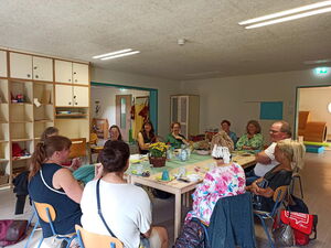 Menschen sitzen rund um einen Tisch in einem Raum. Die Menschen im Vordergrund sind nur mit dem Rücken zu sehnen. Sie sind im Gespräch.