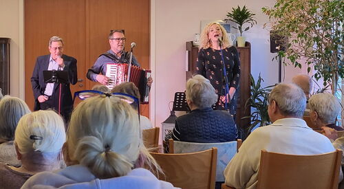 Ein Konzert. Bewohner sintzen auf Stühlen. Die Musiker und eine Sängerin stehen für dem Publikum.